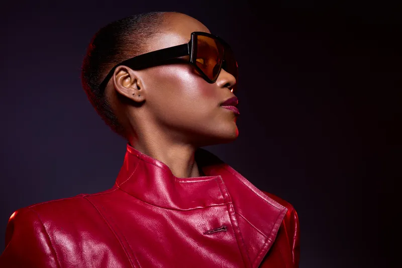 Stylish African American woman with dark skin wearing large sunglasses and red leather jacket looks confidently to the side on dark background. Concept of fashion, elegance, and modern style.