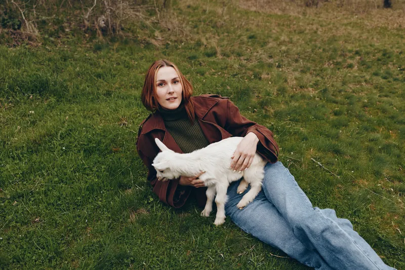 Relaxed woman lies on grass with a playful goat kid beside her, capturing countryside vibes and gentle companionship. Casual fashion, cozy mood, and natural lighting convey a wholesome lifestyle
