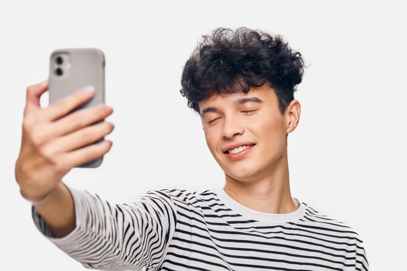 young man taking selfie with smartphone, smiling happily with closed eyes, casual striped shirt, bright studio background, natural lighting