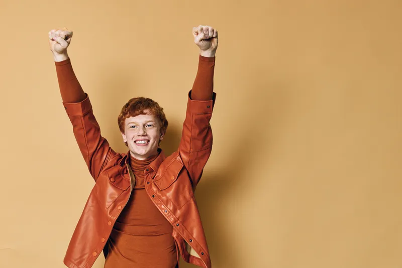 Young man with red hair and freckles wearing orange jacket and turtleneck raising arms celebrating success joy happiness isolated on beige background smiling face