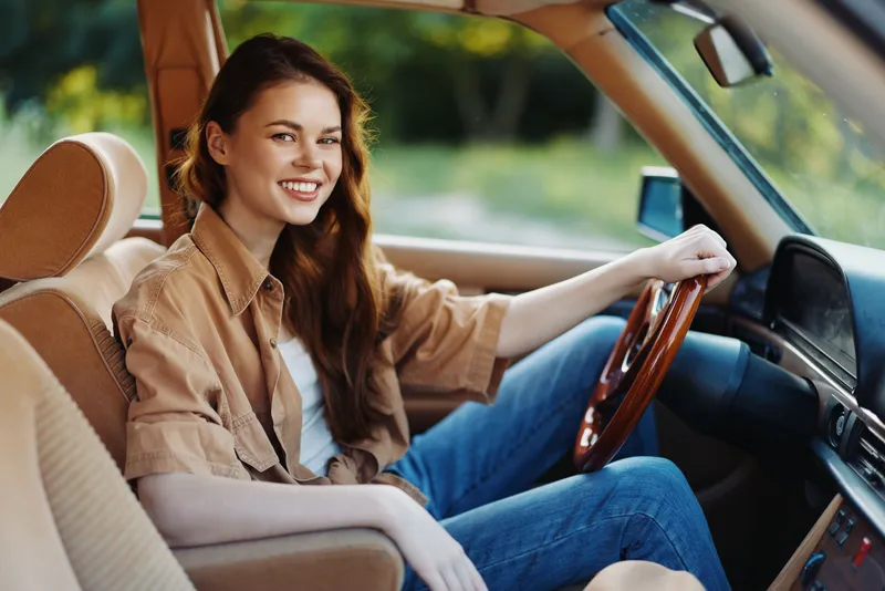 Young woman smiling joyfully in a stylish car, wearing a casual outfit with a light brown shirt, jeans, surrounded by a natural green background.