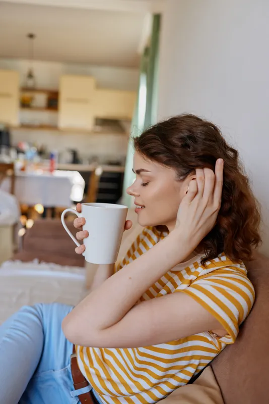 Relaxed woman with striped shirt sipping coffee at home, seated on a sofa, soft natural light, cozy lifestyle moment, casual comfort, peaceful morning mood.