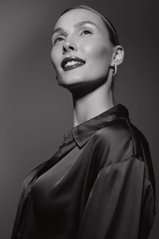 Confident white woman with fair skin and slicked back hair smiles in a black satin shirt against a dark background.