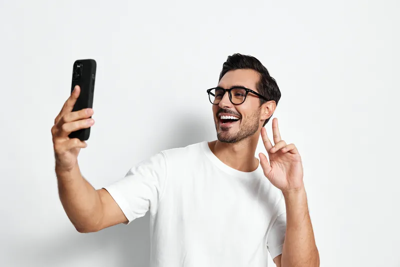 Happy smiling man wearing glasses takes a selfie with smartphone, showing a peace sign isolated on plain light background. Joyful lifestyle concept.