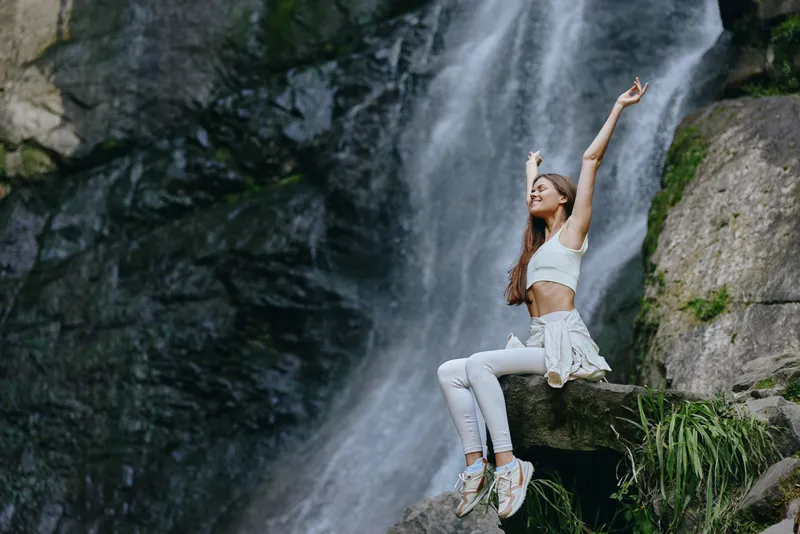 Young woman enjoying nature near waterfall, wearing white activewear, sitting on rock with arms raised, expressing freedom and happiness outdoors in a lush green forest.