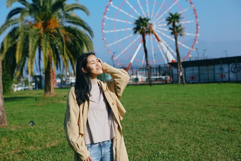 Outdoor woman in a casual coat enjoys the park breeze with a ferris wheel in the background, capturing a serene mood, bright day, green grass, and a fashion inspired moment.