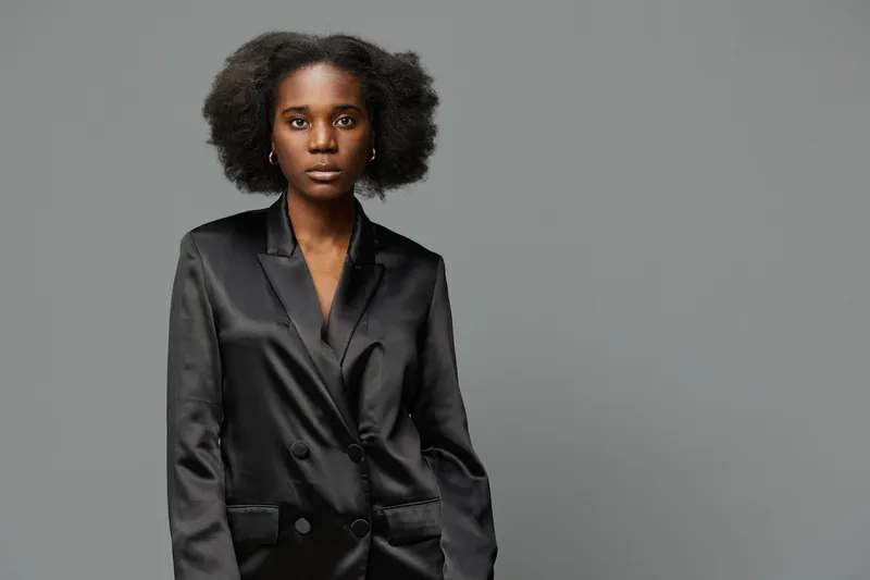 Elegant black woman in a stylish satin blazer, showcasing confident emotion against a neutral gray background, perfect for fashion and professionalism