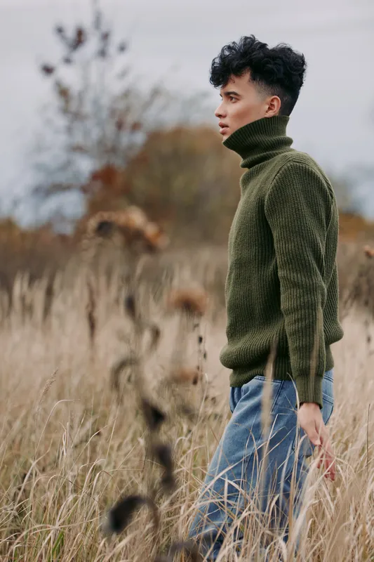 Young man in a green turtleneck walks through tall dry grass, a candid portrait capturing authenticity and quiet reflection with natural lighting and tactile sweater texture.