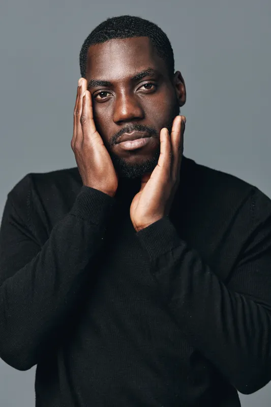 African man with dark skin in black sweater posing with hands on face in creative conceptual visual style. Portrait, serious expression and studio lighting for artistic photography series.