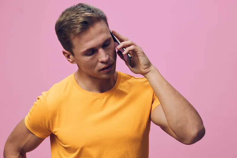 Man talking on mobile phone with thoughtful expression wearing casual yellow t-shirt against pink background. Studio portrait of a young adult male communicating and listening. People lifestyle concep