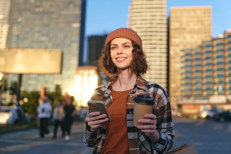 Woman in city holding coffee and phone, smile lit by golden hour glow, candid authenticity capturing mindful living and emotional storytelling in a warm urban lifestyle portrait with relaxed