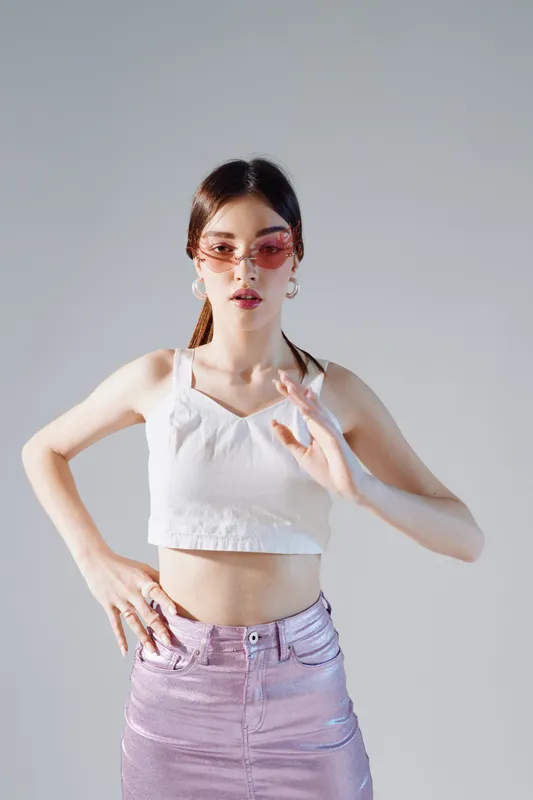 Young woman in stylish pink skirt and white top poses confidently wearing pink sunglasses. Studio shot on grey background. Concept of woman beauty fashion and modern style.