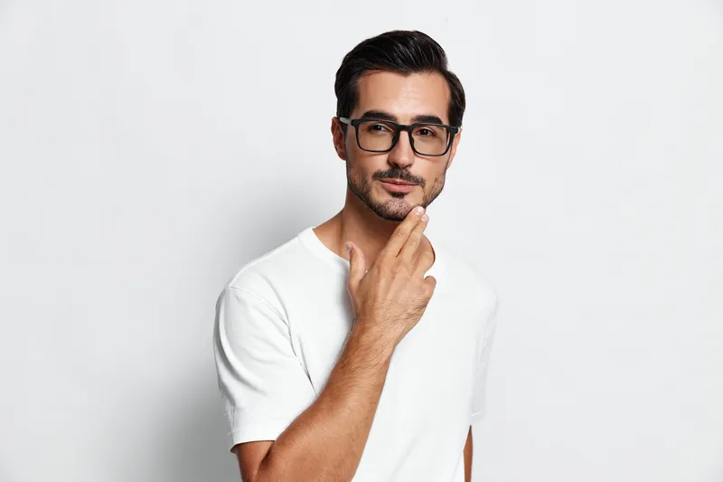 Thoughtful young man wearing glasses, posing confidently with hand on chin, isolated on plain solid background. Modern lifestyle and self-reflection concept.