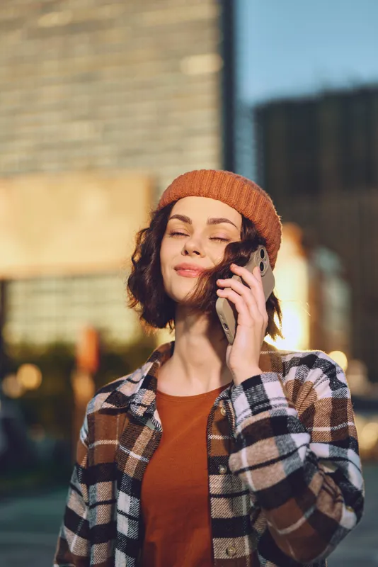 woman on phone in urban street smiling with beanie and sunlight, candid autumn portrait capturing authenticity and mindful living with golden hour glow and emotional storytelling mood.