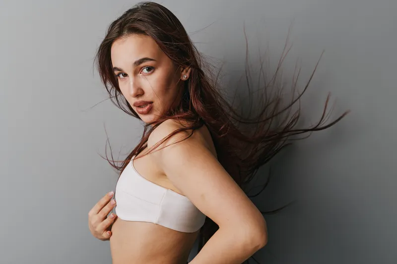 Young woman with long hair wearing white top posing confidently against neutral background. Female model looking at camera with natural makeup and flowing hair in studio portrait.