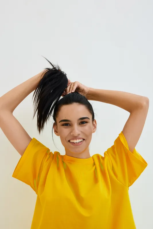 Smiling woman in yellow shirt lifting her hair with both hands, joyful expression, casual style, bright clothing, isolated background, youthful energy