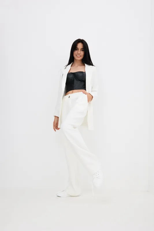 Smiling woman in white suit poses confidently against plain white background wearing stylish black top and wide-leg trousers, showcasing modern fashion and casual elegance.