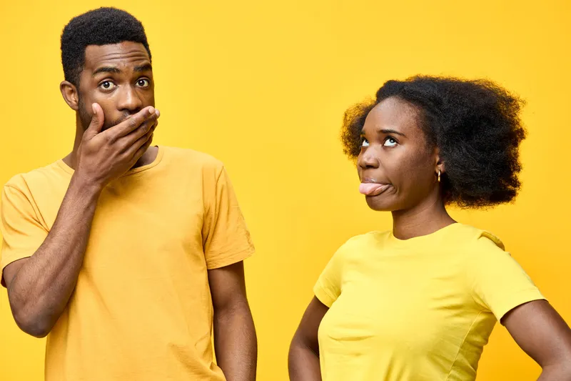 A playful couple in matching yellow shirts express fun emotions, with one covering their mouth and the other sticking out their tongue against a bright yellow background