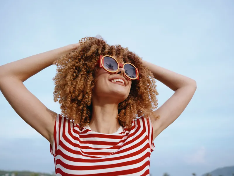Young woman with curly hair and round sunglasses smiles under a clear blue sky, wearing a red and white striped tee, radiating carefree summer energy.