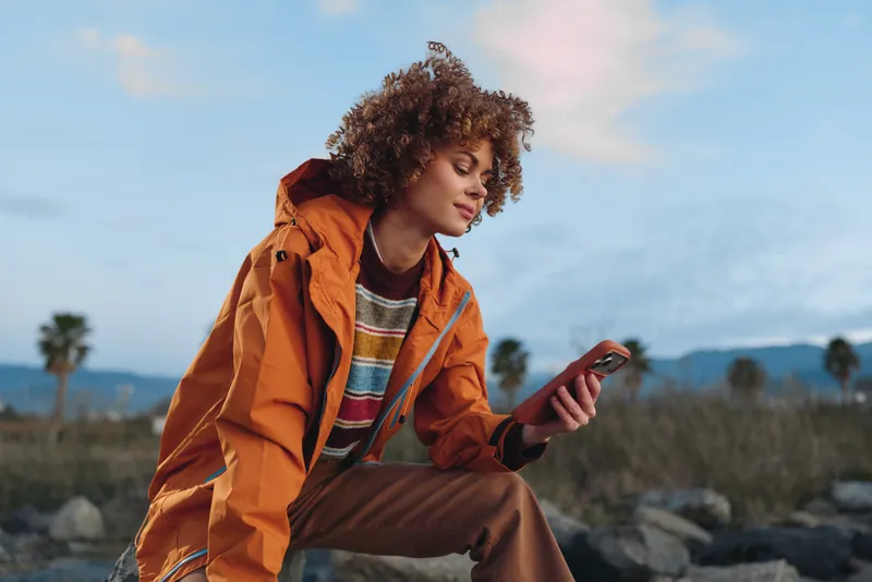 Woman lifestyle outdoors, smartphone in hand, orange jacket, curly hair in light breeze, sitting on rocks, looking at phone, relaxed smile, landscape horizon, soft daylight