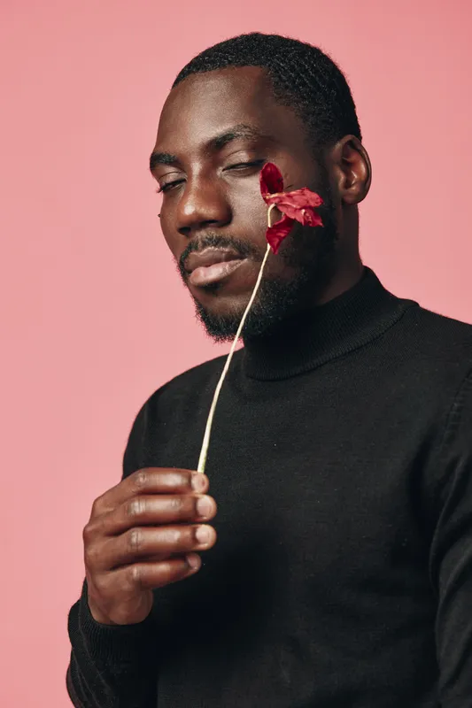 African man with closed eyes holding red flower near face in creative conceptual visual style. Portrait of thoughtful black male in black turtleneck against pink background for artistic expression.