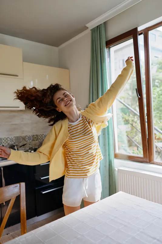 A young woman in a striped shirt and yellow jacket dances in a bright kitchen by a large window, arms raised, hair swinging, conveying carefree home energy and joyful morning vibes.