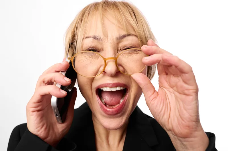 Elderly woman with glasses laughing on phone, communication moment, closeup portrait on white background, blonde hair, hands adjusting eyeglasses, joyful expression, bright