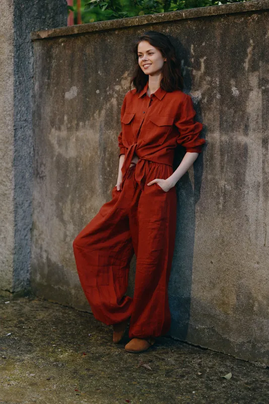 A stylish woman in a rust jumpsuit leans against a concrete wall, enjoying a casual outdoor moment. A confident, relaxed lifestyle moment for fashion storytelling