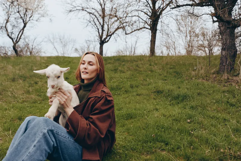 A woman sits on the grass in a park, holding a goat kid with a gentle smile, capturing a calm outdoor lifestyle moment in nature and animal company.