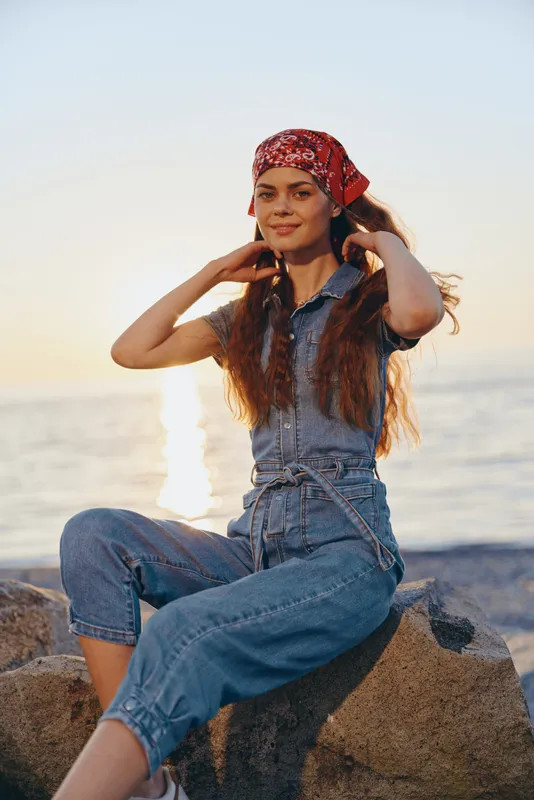 Lifestyle woman in modern boho-western denim street style sitting by the sea wearing red bandana, enjoying golden hour sunlight with warm film color tones and relaxed mood.