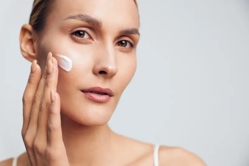 Young woman with clear skin applies moisturizer cream on her cheek while looking thoughtfully at the camera in a bright studio setting.