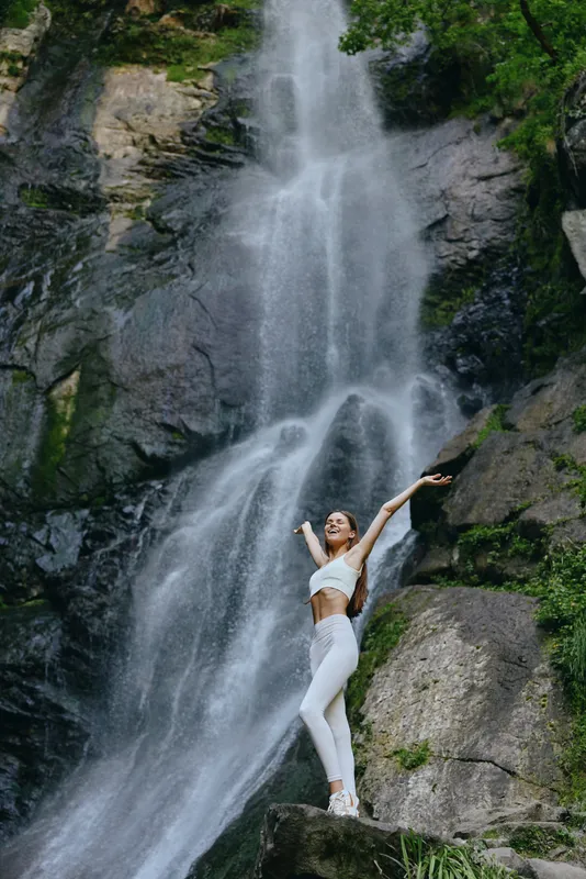 happy woman standing near waterfall in white activewear enjoying nature surrounded by green forest and rocks, celebrating outdoor adventure and freedom