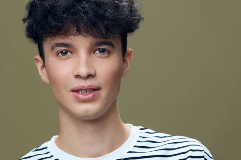 young man with curly hair wearing striped shirt looks confidently into camera isolated on olive background portrait of casual stylish man with short haircut