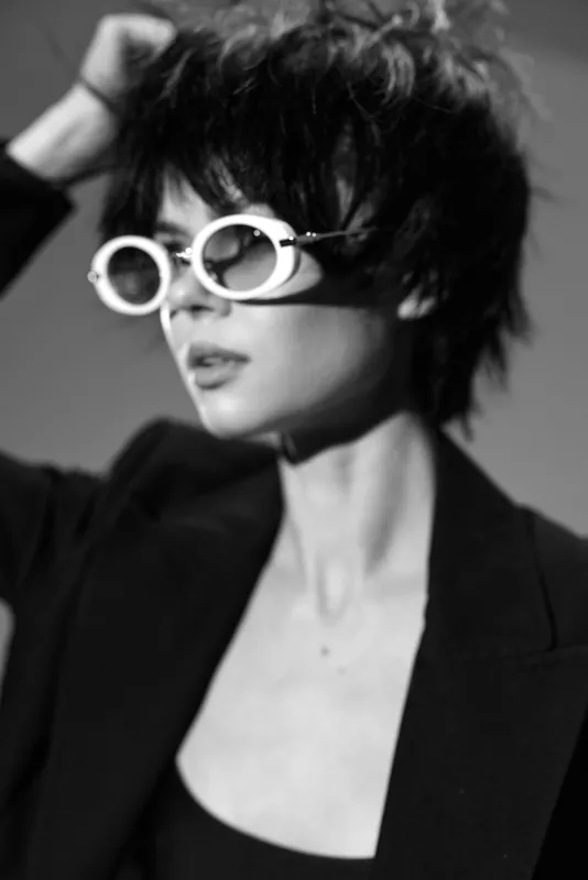 Black and white portrait of a stylish woman wearing round sunglasses and a blazer with a modern haircut and serious expression.