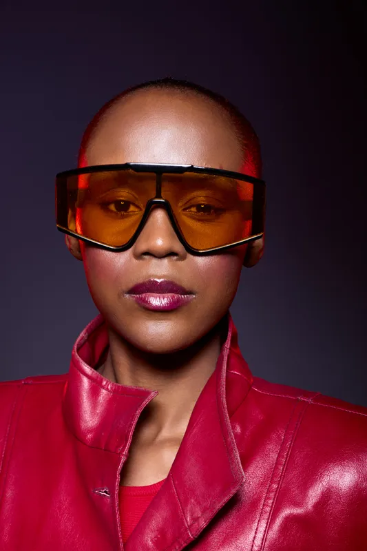 Close-up portrait of a confident African woman with dark skin wearing large orange sunglasses and a red leather jacket. Studio lighting highlights her smooth complexion and bold fashion style.