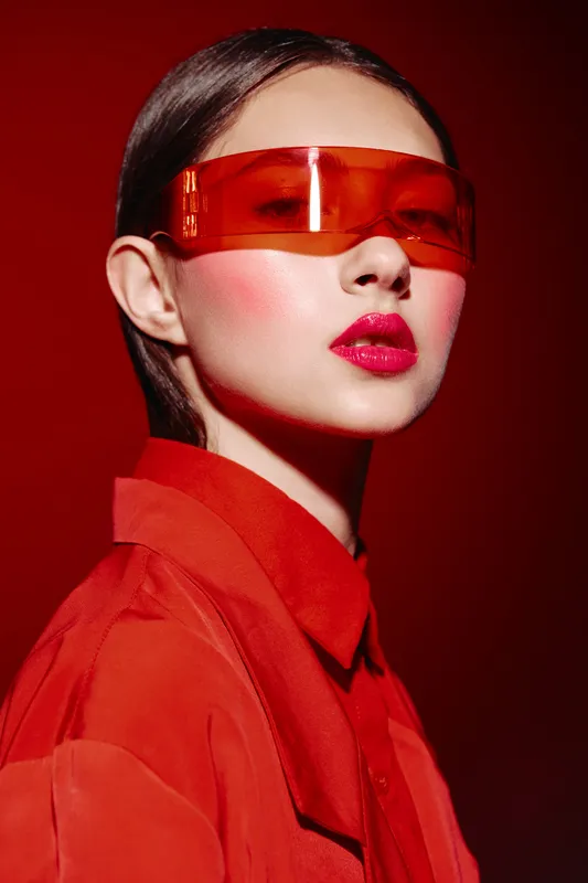 Young woman in red jacket and bold red lipstick wearing large orange translucent fashion glasses posing against bright red background. Concept of beauty, fashion, and creative style.