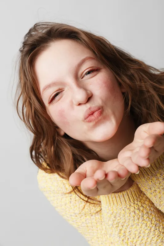 Young woman with wavy hair wearing yellow sweater blows a kiss with closed eyes. Studio portrait on white background. Concept of affection, friendliness, and positive emotions.