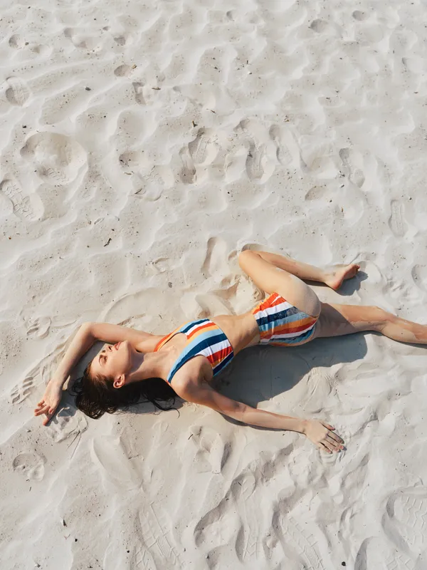 Tranquil scene features a woman lying on bright sand, wearing a striped onepiece swimsuit. Sunlit, relaxed pose conveys carefree summer leisure and warm youthful energy.