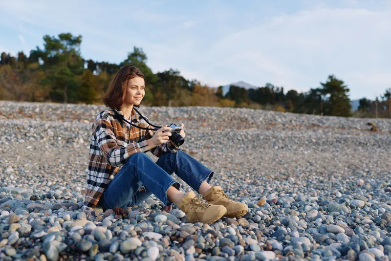 Woman sitting on a pebble beach with a camera, enjoying outdoor photography in a natural setting, casual outfit, daylight and calm mood while capturing scenes