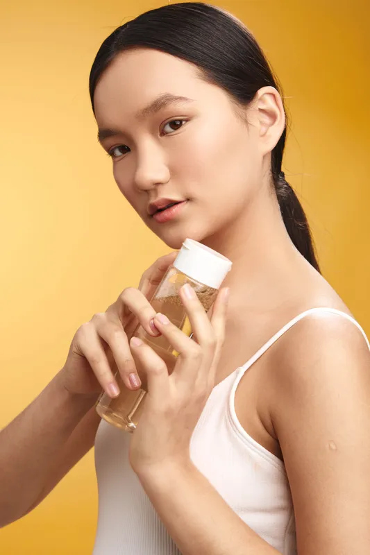 Asian woman in a white tank top holds a clear skincare bottle against a warm yellow backdrop, presenting a confident, fresh look and a sense of personal care.