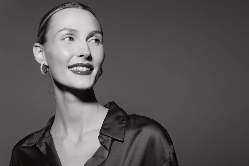 Young white woman with pale skin and light hair smiles gently, dressed in a satin shirt in a studio. Portrait in black and white with soft lighting highlights facial features and expression.