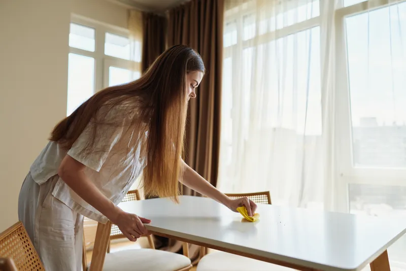 Woman cleaning table at home in bright morning light, wiping dining surface with cloth. Domestic chore and tidy household routine, casual outfit and focused care in living room interior