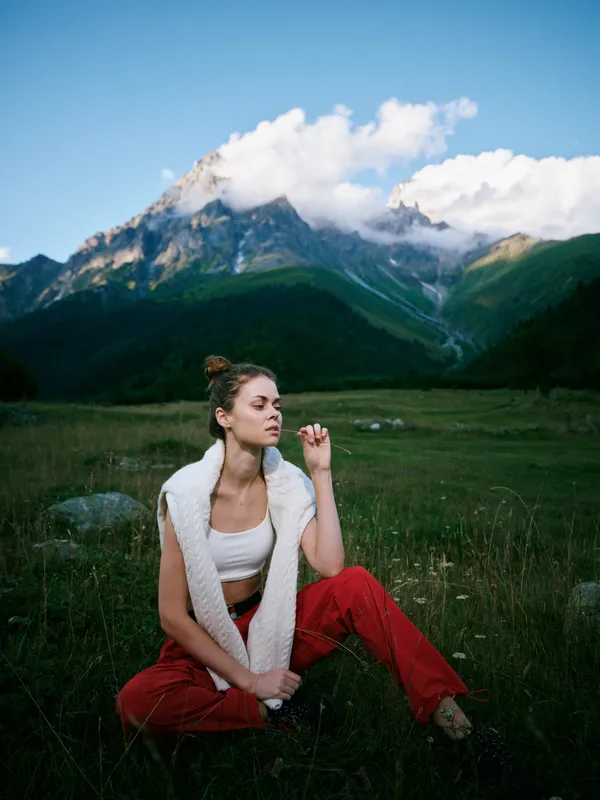 Woman sits in a meadow with red pants and white vest, towering mountains and blue sky behind, outdoor fashion and serene alpine scenery for lifestyle and travel use