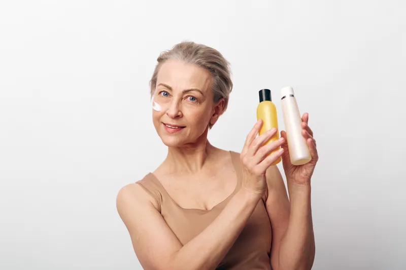 Mature woman with natural skin holds two cosmetic bottles, smiling gently. Skincare, beauty, and wellness concept focused on aging care and natural facial treatment on white background.