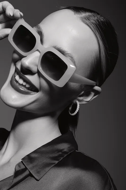 Young caucasian woman with fair skin wearing large sunglasses and hoop earrings smiles confidently. Black and white studio portrait with elegant satin shirt. Concept of fashion and style.