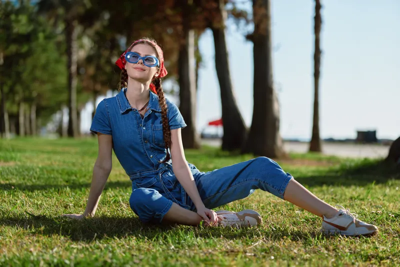 Lifestyle woman in modern boho-western denim street style sitting on grass outdoors with film color tones, enjoying sunny day and relaxed mood in stylish casual outfit.