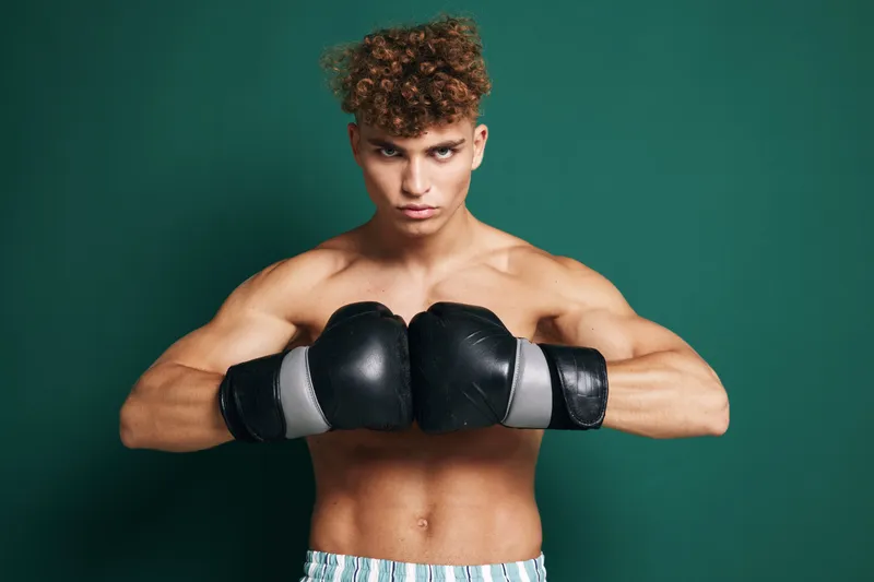 Young male boxer with curly hair wearing black gloves and striped shorts posing shirtless in front of green background for fitness and sports training concept.
