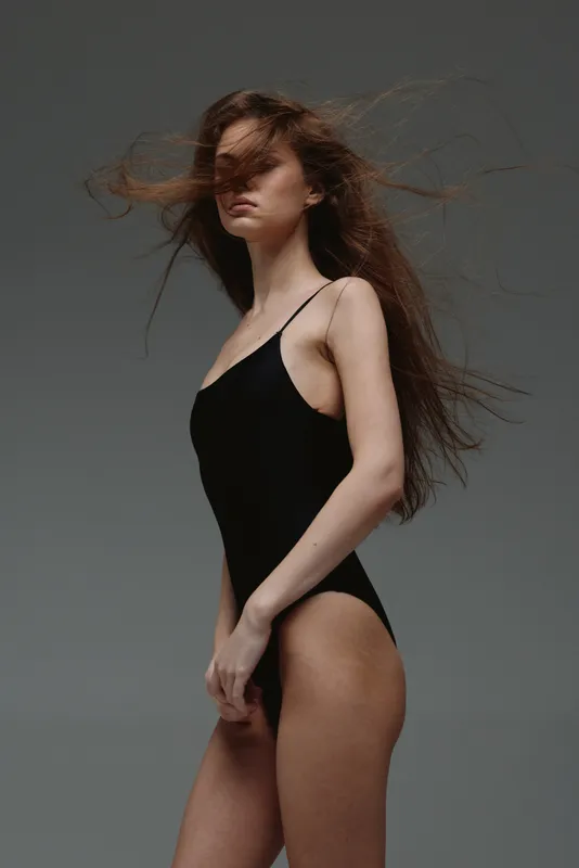 Young woman in black swimsuit with windswept hair standing against gray background in studio setting