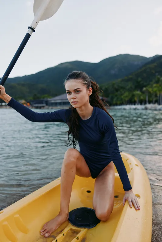 Young woman kneels on a yellow kayak near the shore, raising a paddle with focus. Wetsuit-clad athlete, calm lake, and distant mountains create a vivid outdoor sport scene.