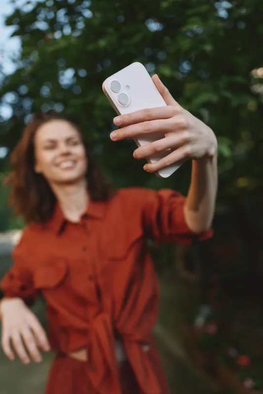 Cheerful woman outdoors, taking a selfie in a red dress, sunny day, natural light, relaxed pose, gentle breeze, vibrant colors, lush greenery background
